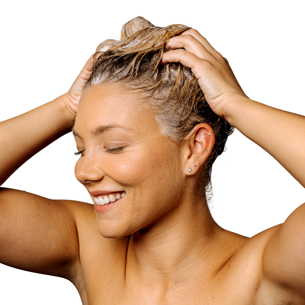 A person smiling with eyes closed, massaging Geologie's Self Care Gift Set into their hair against a black background.
