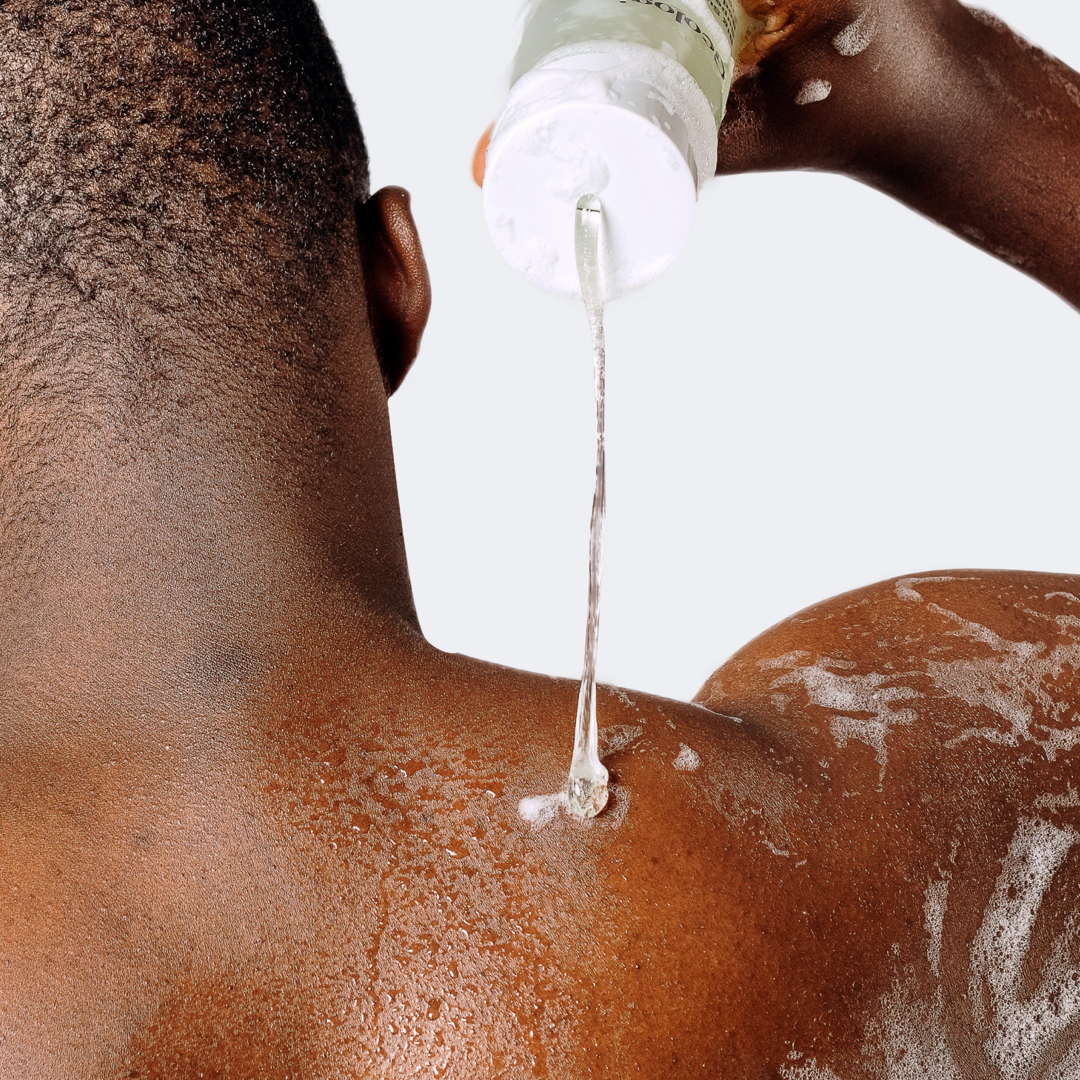 A person pours Geologie's Acne Control Superclean Body Wash onto their shoulder while taking a shower. The gel is flowing from a bottle onto their wet, soapy skin, helping with breakouts prevention.