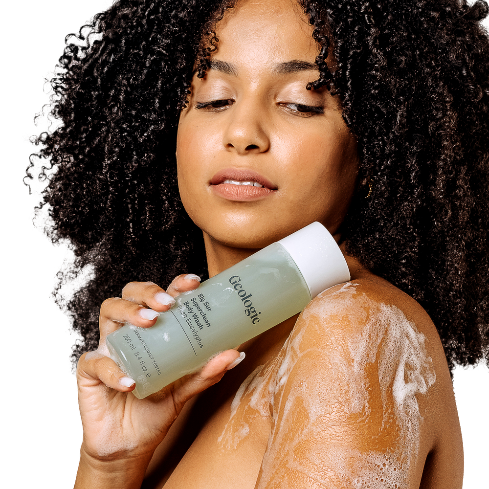 A woman with curly hair holding a power-packed Geologie Self Care Gift Set, with foam on her shoulder.