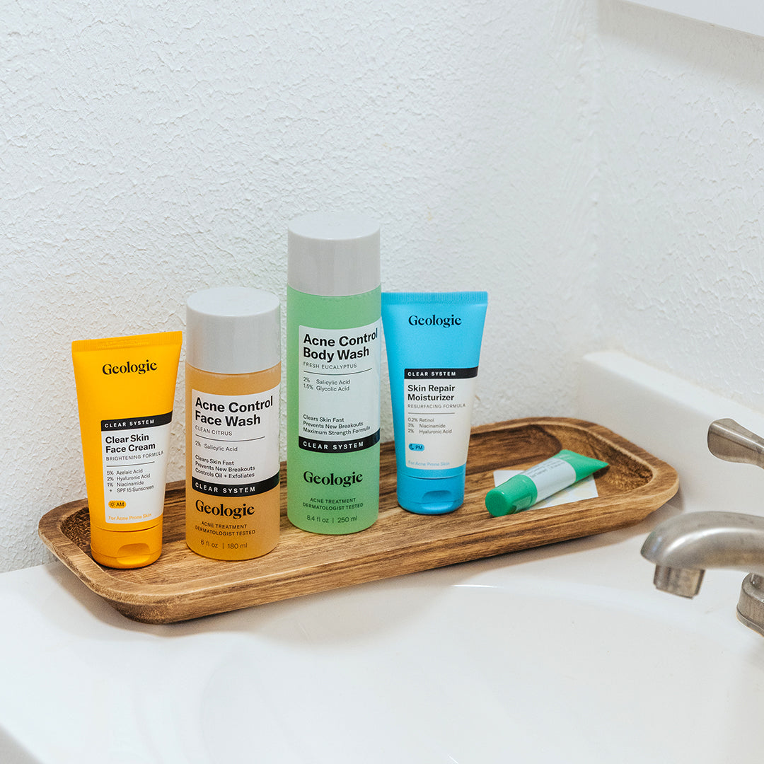 The Geologie Clear System Set, featuring face wash, body wash, moisturizer, and face cream, is neatly arranged on a wooden tray beside a sink.