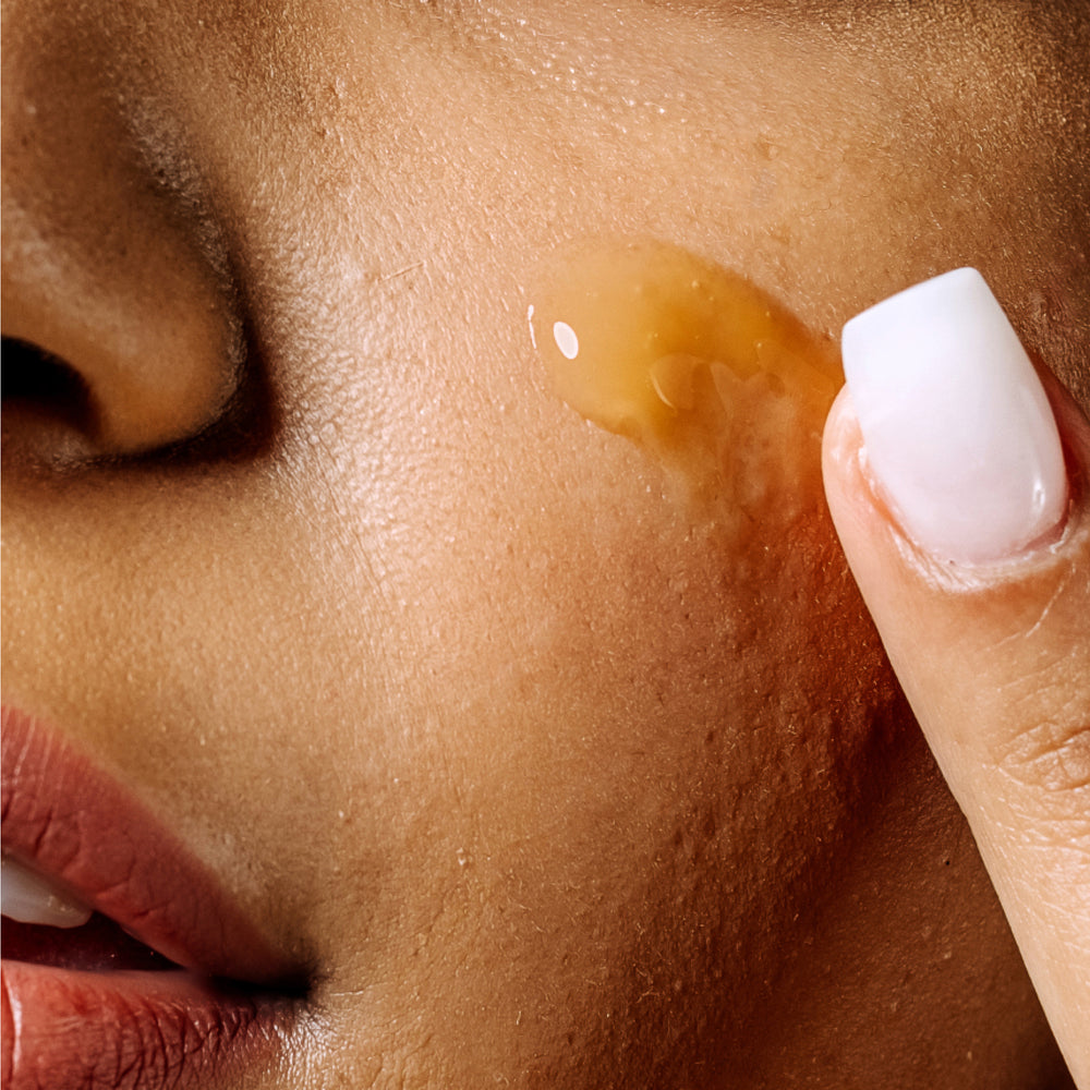 Close-up of a person applying Geologie's Vitamin C+E Ferulic Serum to cheek, focusing on skin and fingertips with a droplet of serum visible.