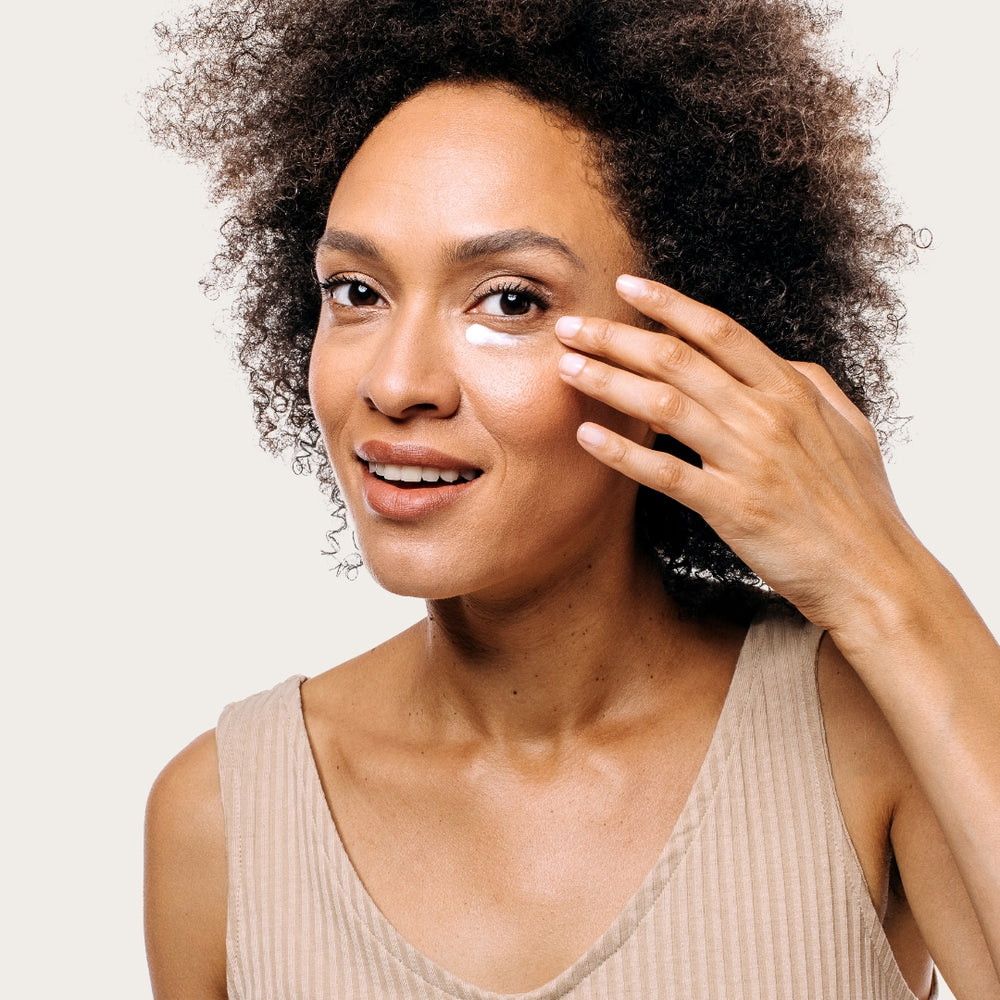 Woman with curly hair touching her face and looking at the camera against a white background, her skin glowing from the Geologie Dark Under Eye Duo.